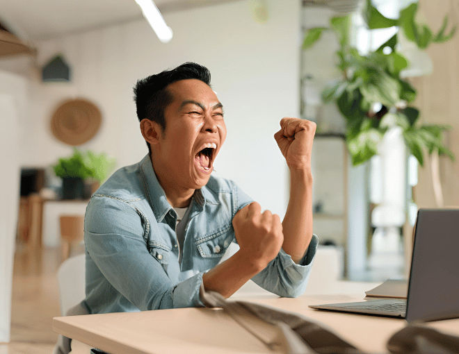 An Indonesian man, seated in a well-lit room, excitedly punches the air with a clenched fist while focusing on his laptop.